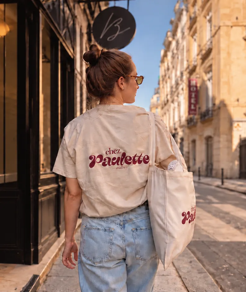 Une femme de dos dans une rue de Paris, devant une devanture noire au soleil, en t-shirt crème Chez Paulette et jean, le tote bag Chez Paulette à l’épaule