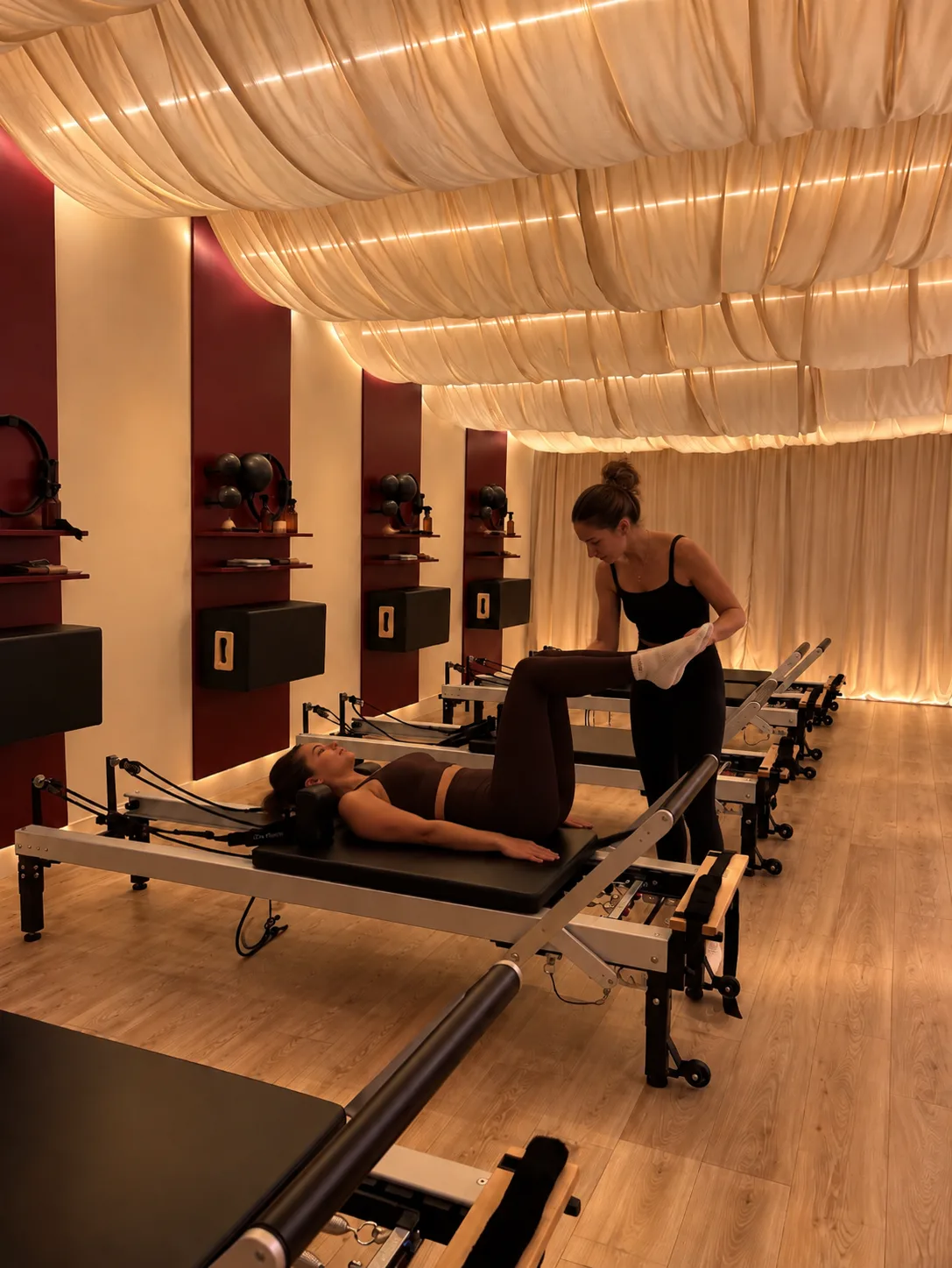 Une coach corrige la jambe d’une élève allongée sur un Reformer, sous le plafond drapé du studio Chez Paulette à Sartrouville